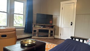 Quarto com televisão sobre suporte de madeira, janela com cortinas azuis, porta e cama com manta azul.
