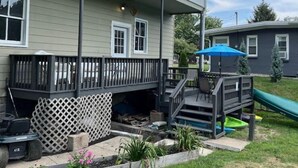 Uma casa de dois andares com um alpendre coberto, um espaço de recreação com escorrega e um jardim cercado.