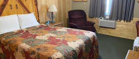 A room with a wooden headboard bed, a quilted bedspread, a chair, a lamp, a wall clock, a bear picture, and a window with curtains.