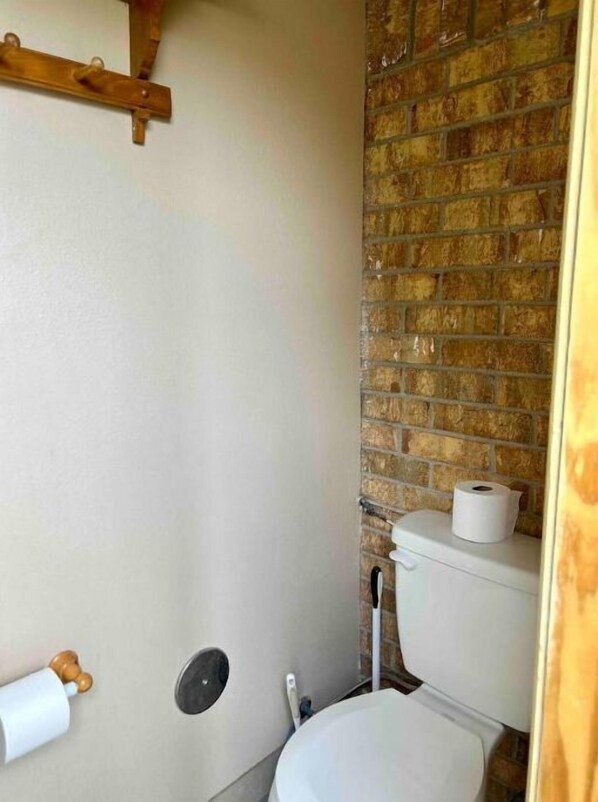 A bathroom with a white toilet, a wooden shelf, and a brick wall.
