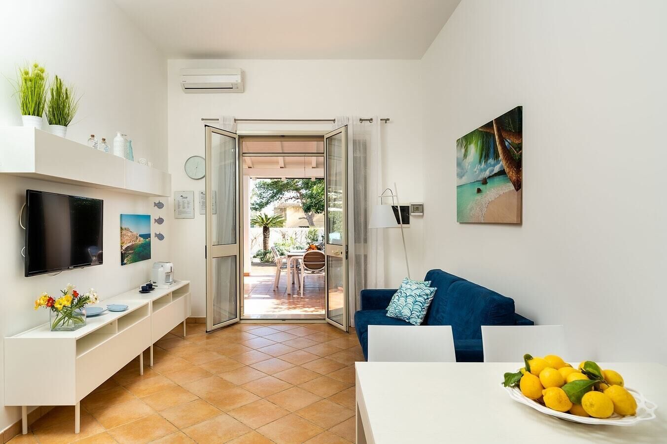 A modern living room with a TV, a dining area with a table and chairs, a sofa with cushions, and a balcony with a view of the outdoors.