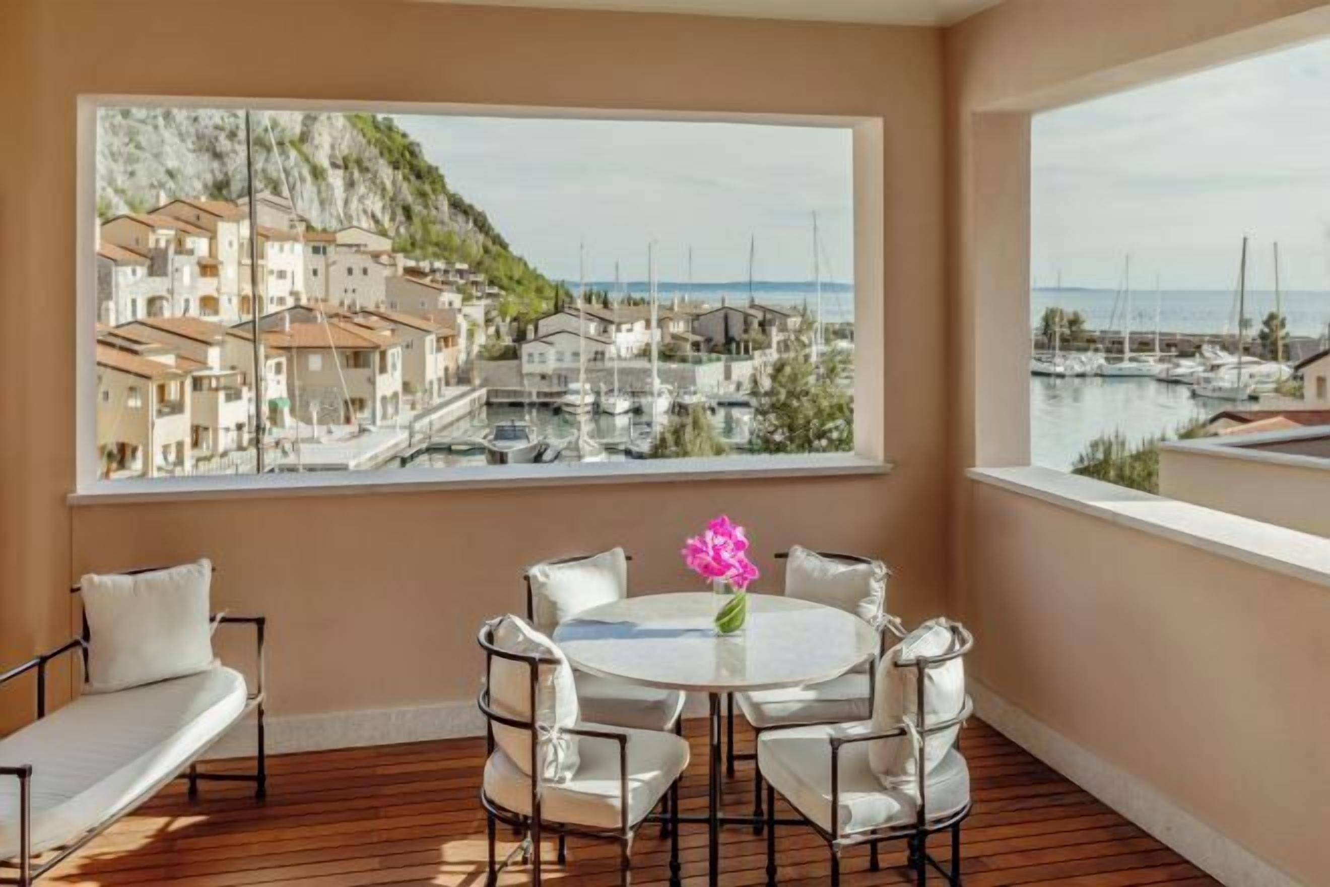 A balcony with a round table and chairs, a sofa, and a large window with a view of a coastal town and marina. - Tivoli Portopiccolo Sistiana Resort (Duino-Aurisina)