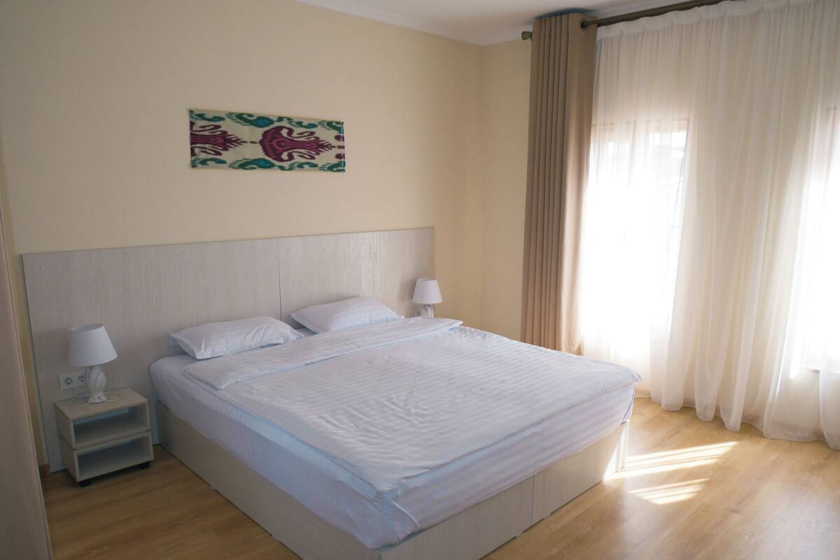 Een netjes opgemaakt bed in een hotelkamer, met een hoofdbord, twee nachtkastjes met lampen en een raam met gordijnen.