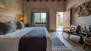 A bedroom with a large bed, a wooden ceiling, a stone wall, a window, and a dining area with a table and chairs.