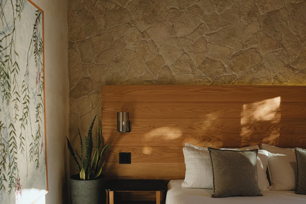 A bedroom with a stone wall, a wooden headboard, a bed with white bedding, a bedside table, a potted plant, and a wall-mounted lamp.