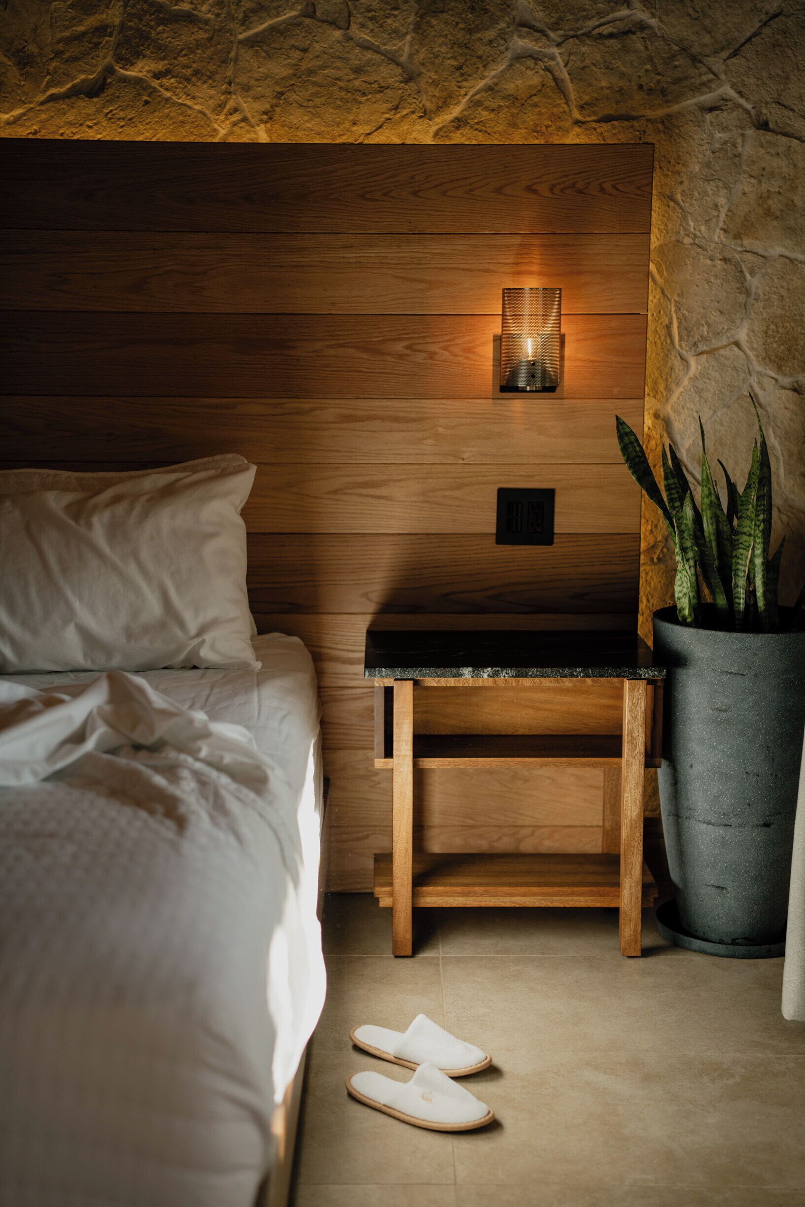 Un dormitorio con cabecera de madera, una mesita de noche con lámpara, una planta en maceta y un par de zapatillas en el suelo.