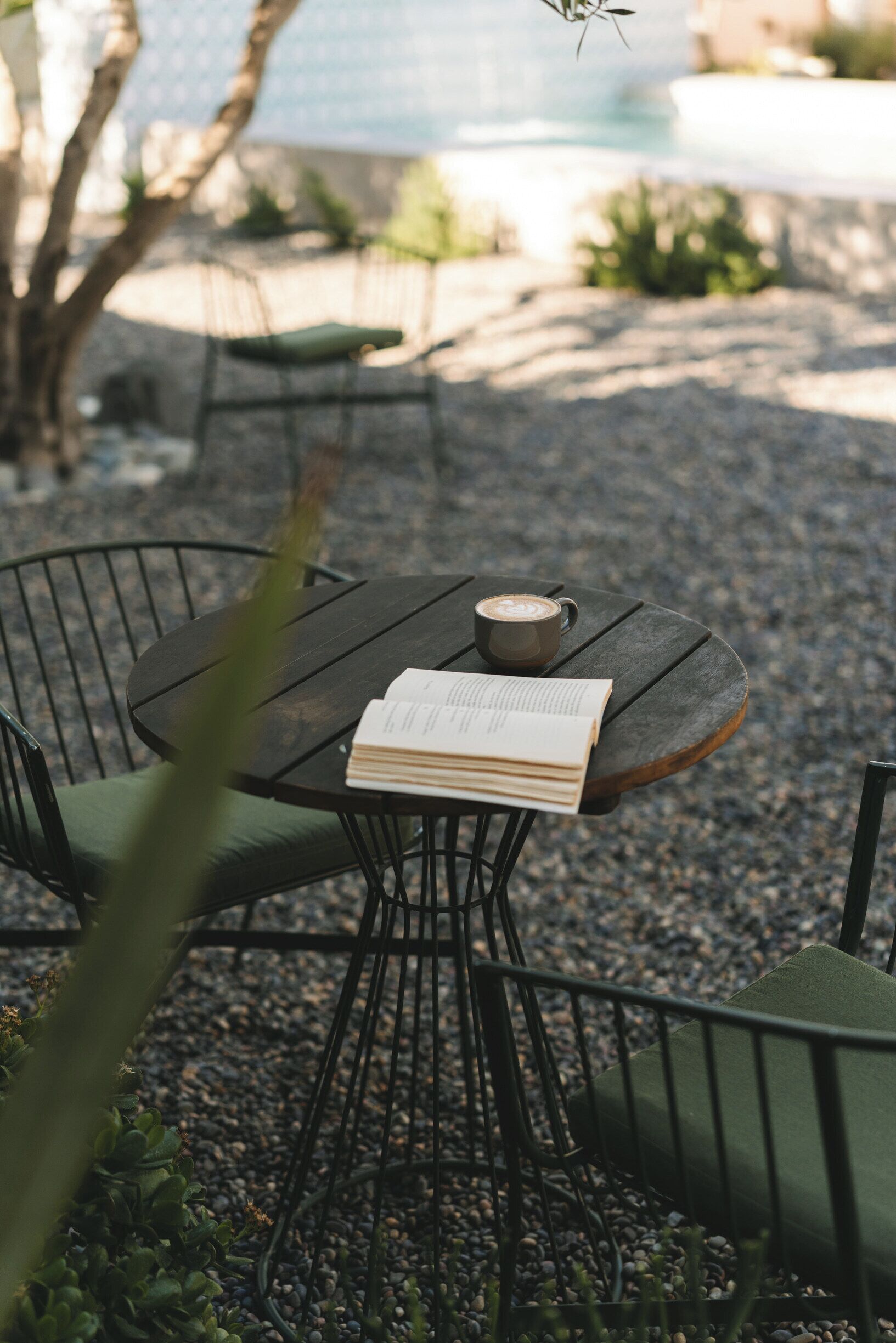 Une table ronde avec un livre et une tasse, entourée de chaises avec des coussins, dans un cadre extérieur agrémenté de gravier et de plantes.