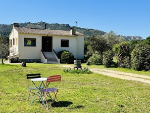 A single-story building with a tiled roof, a front door, and a small set of stairs leading to the entrance. There is a grassy area in front with a table and chairs. - Can Bagudanch CostaBravaSi - Pool, A/C, Golf (Santa Cristina d'Aro)