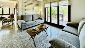 A spacious living room with a grey sofa, a wooden coffee table, and a balcony with a view of greenery.