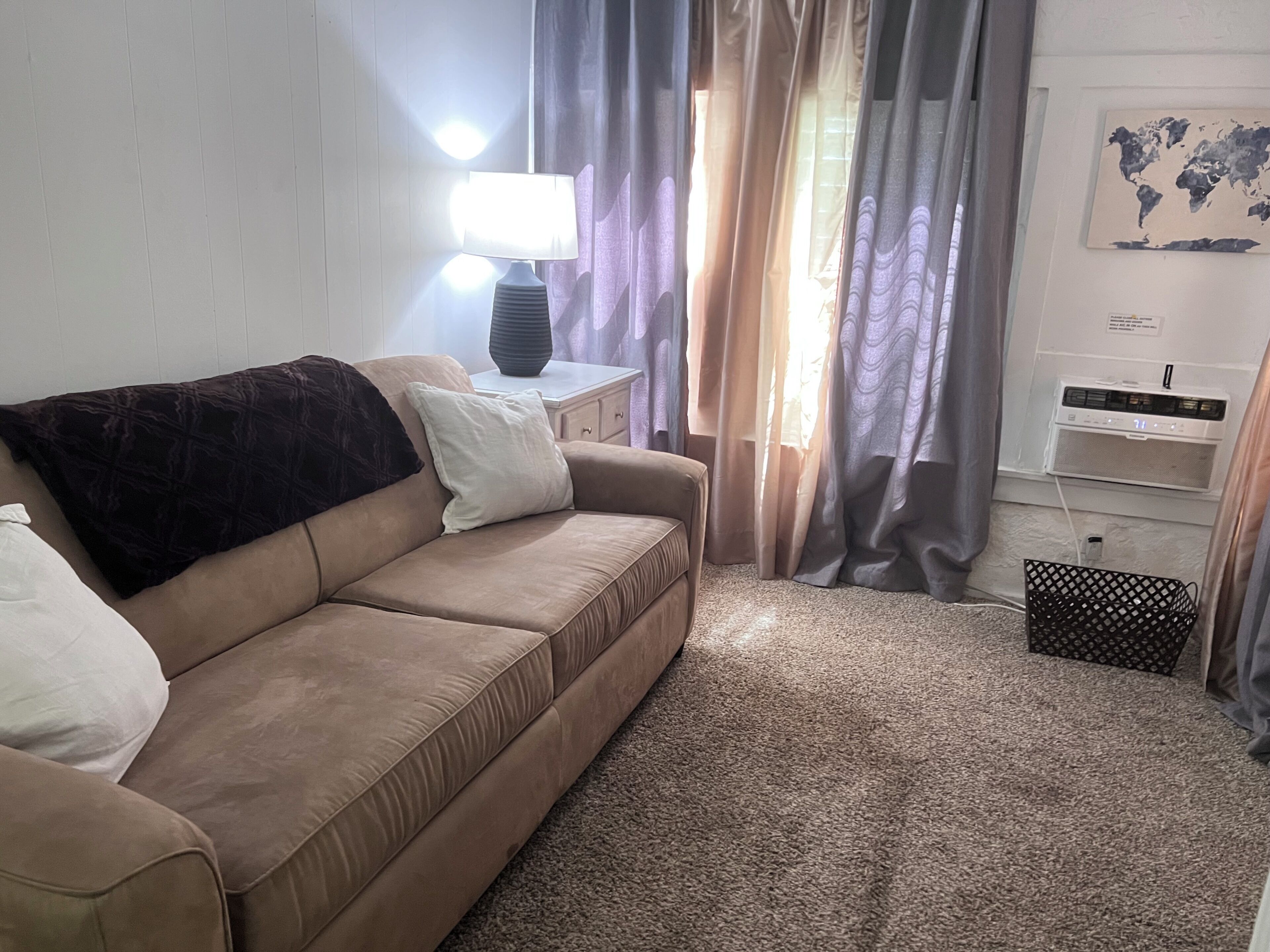 A living room with a beige sofa, a side table with a lamp, a world map on the wall, and a small black basket.