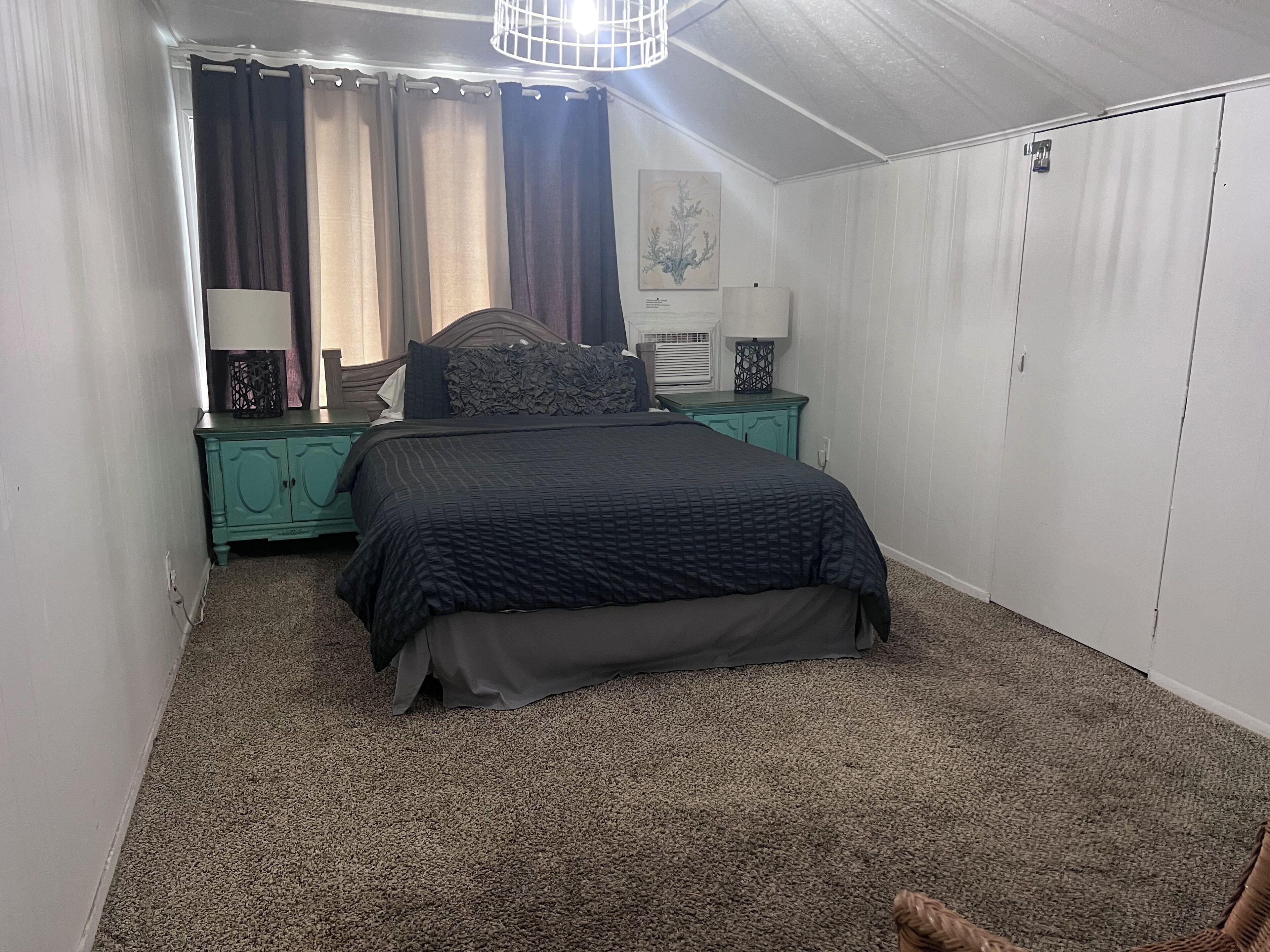 A bedroom with a bed, a nightstand, a dresser, a window with curtains, and a wall-mounted light fixture.