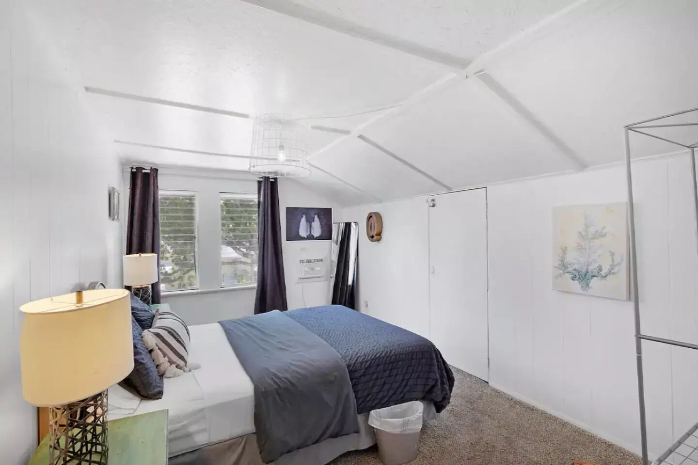 Une chambre à coucher avec un lit, une lampe de chevet, une fenêtre avec des rideaux, un climatiseur fixé au mur, une porte et un tableau.