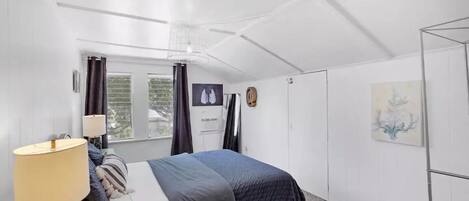 A bedroom with a bed, a bedside lamp, a window with curtains, a wall-mounted air conditioner, a door, and a painting.