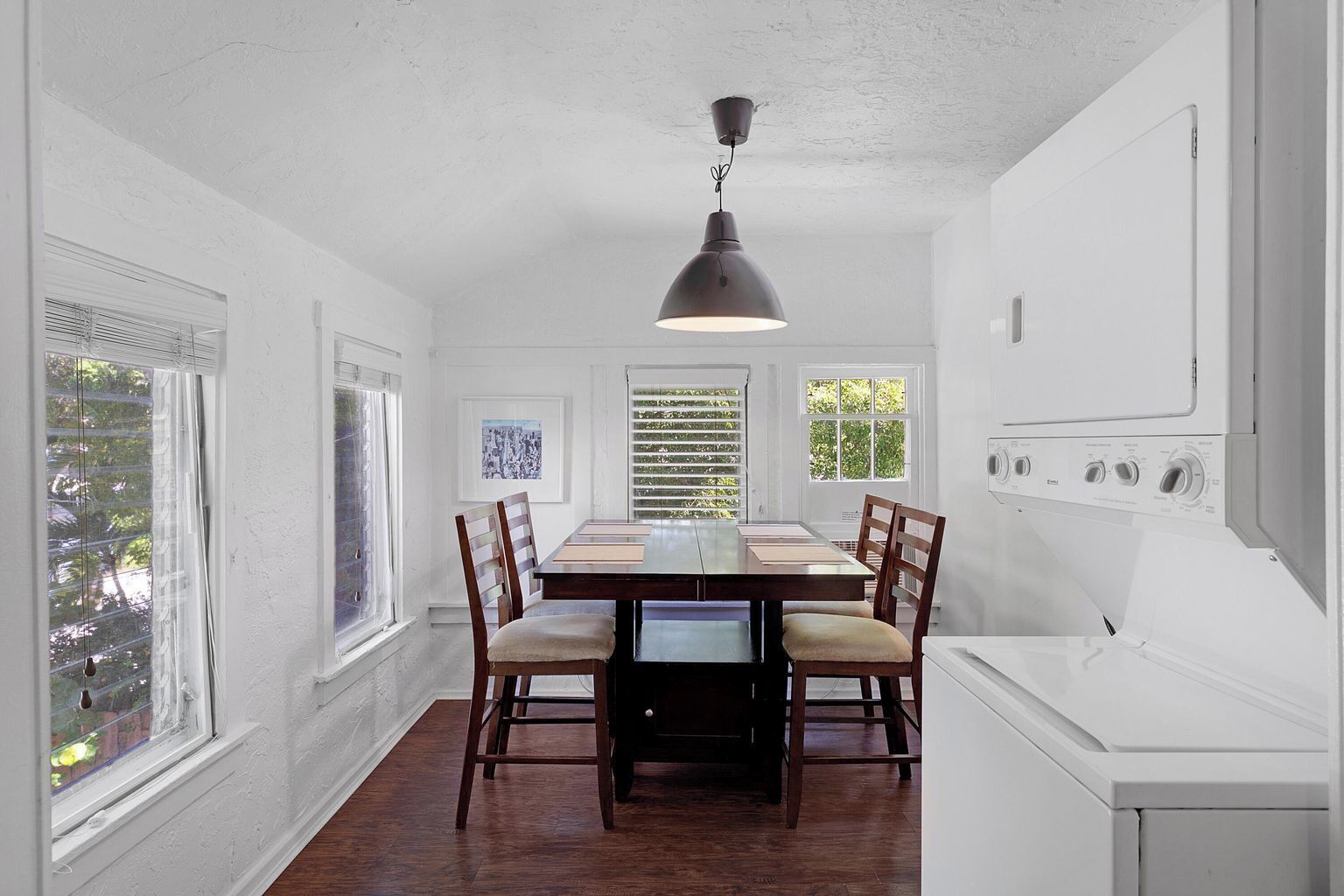 Un coin repas avec une table en bois, des chaises et un luminaire suspendu. On aperçoit des fenêtres avec des stores, un mur blanc et un lave-linge à l’arrière-plan.