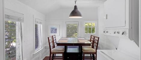 A dining area with a wooden table, chairs, and a pendant light. There are windows with blinds, a white wall, and a washing machine in the background.
