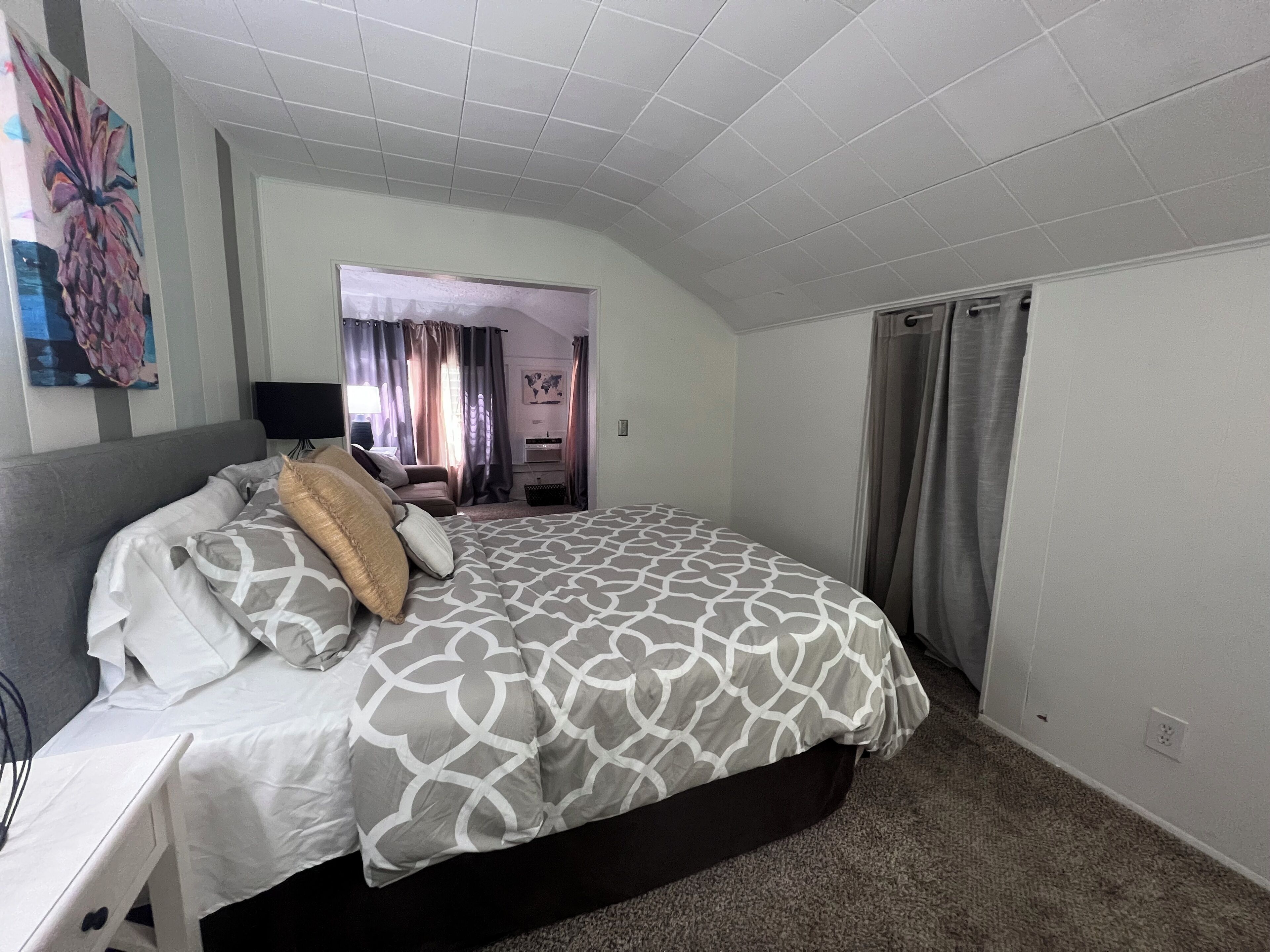 A bedroom with a large bed, a desk, and a chair. There is a window with curtains, a painting on the wall, and a door leading to another room.