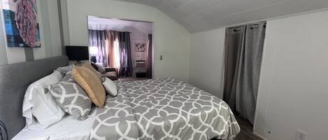 A bedroom with a large bed, a desk, and a chair. There is a window with curtains, a painting on the wall, and a door leading to another room.