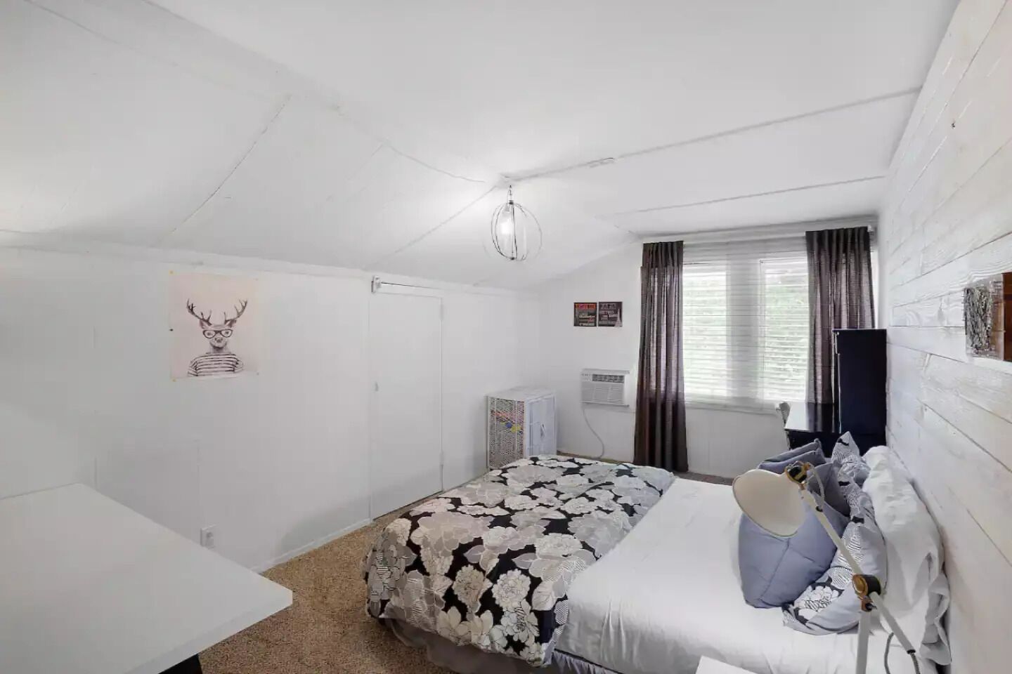 A bedroom with a bed, a desk, a chair, a window with curtains, and a wall-mounted air conditioner.