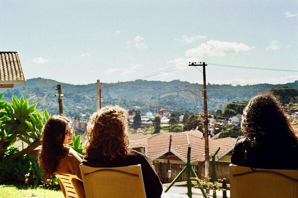 Three individuals seated outdoors with a view of a hilly landscape and a village in the distance. - Mountain chalet - Recanto da Marina (Jardim Primavera Campos do Jordão)