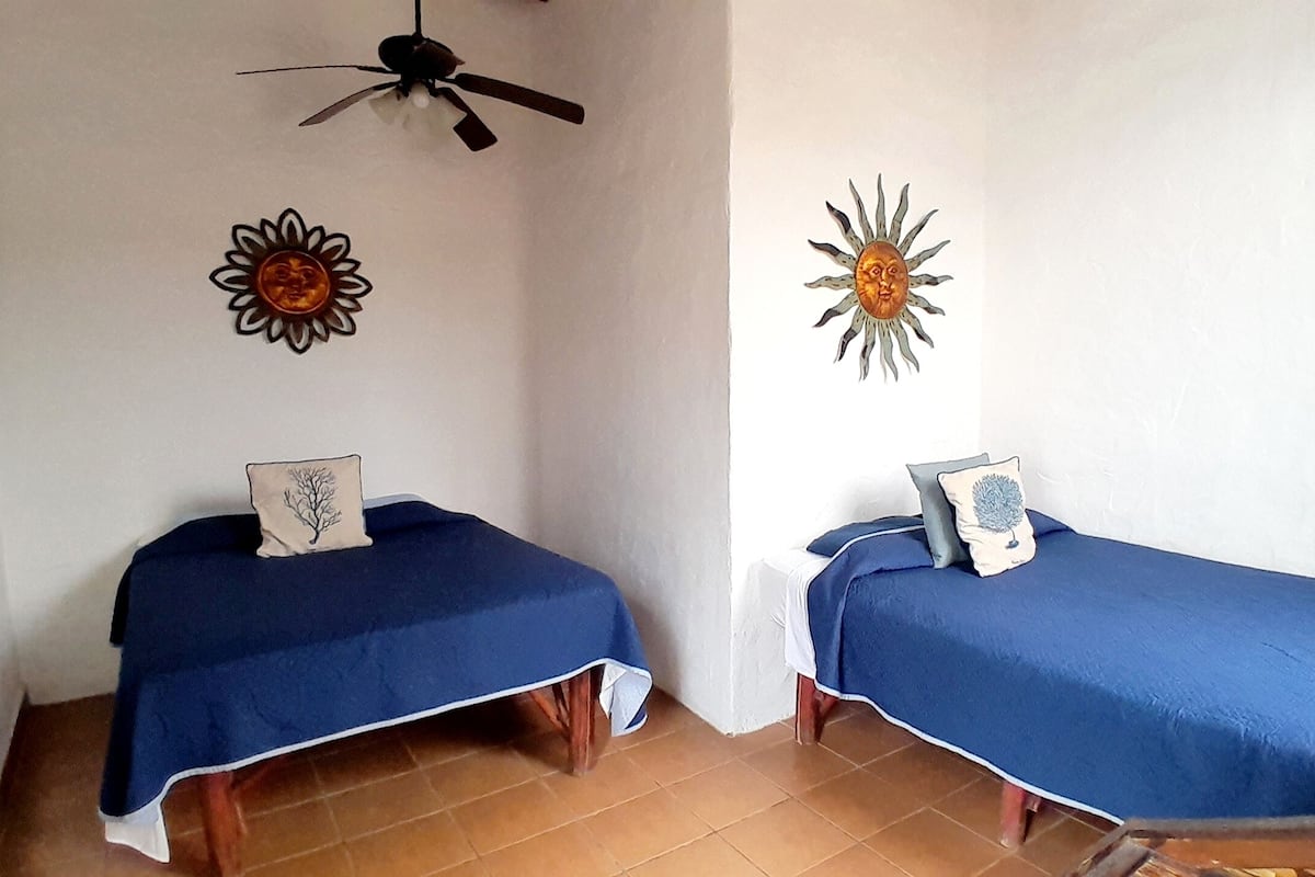 Two single beds with blue covers, a ceiling fan, and decorative sun motifs on the walls.