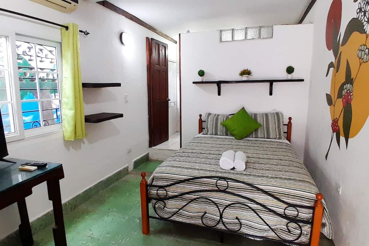 A bedroom with a bed, a TV, a desk, a window with curtains, and a wall decoration.