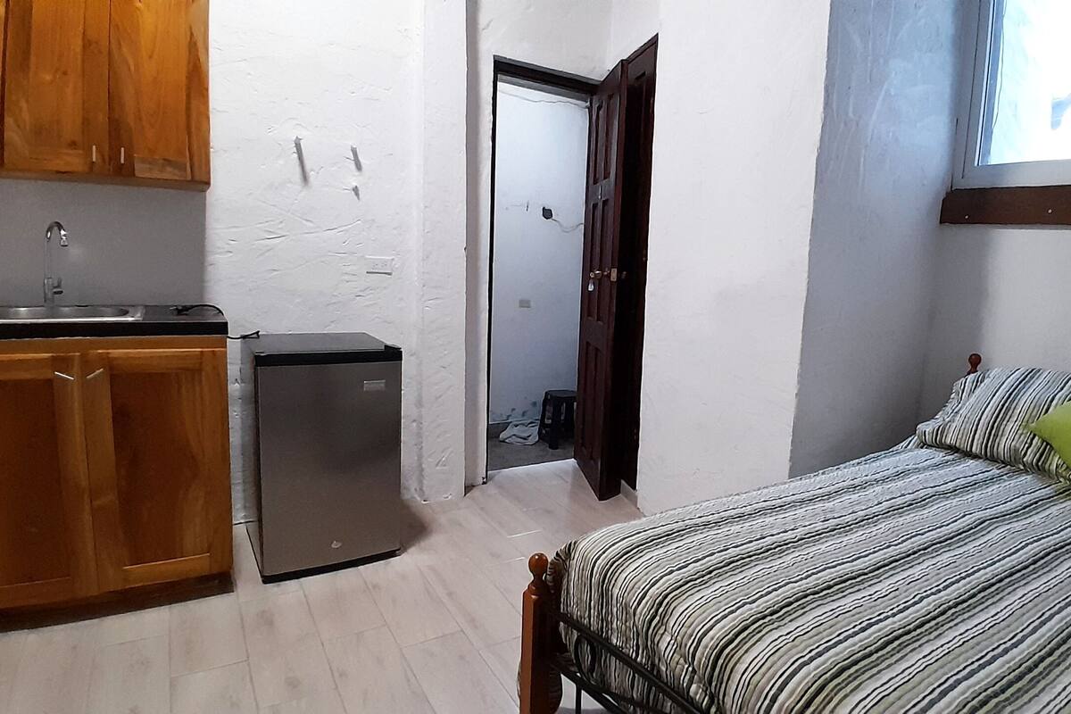 A compact room with a bed, a small kitchenette, and a door leading to another room.