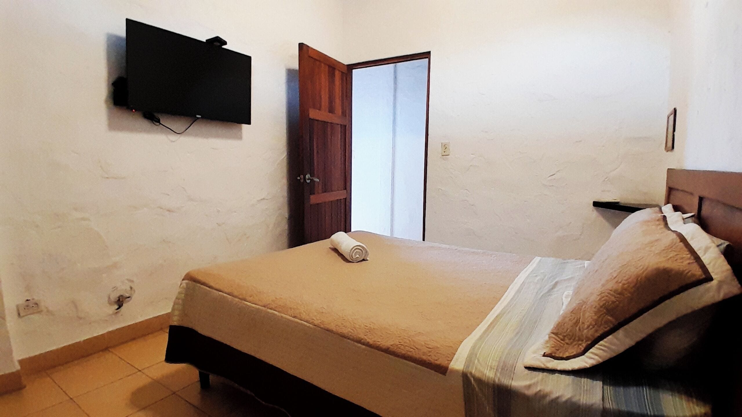 A bedroom with a bed, a TV mounted on the wall, a wooden door, and a small table with a pillow.