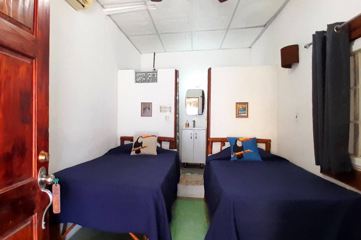 A room with two beds, a wooden door with a number, a small kitchenette, and a window with curtains.