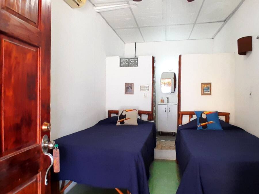 A room with two beds, a wooden door with a number, a small kitchenette, and a window with curtains.
