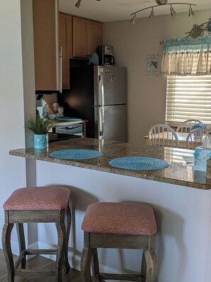 A kitchen with a refrigerator, a coffee maker, and a potted plant. There are two stools and a window with blinds. - Bridget's Beach Retreat, On the beach with views on balcony,bedroom,living room (New Smyrna Beach)