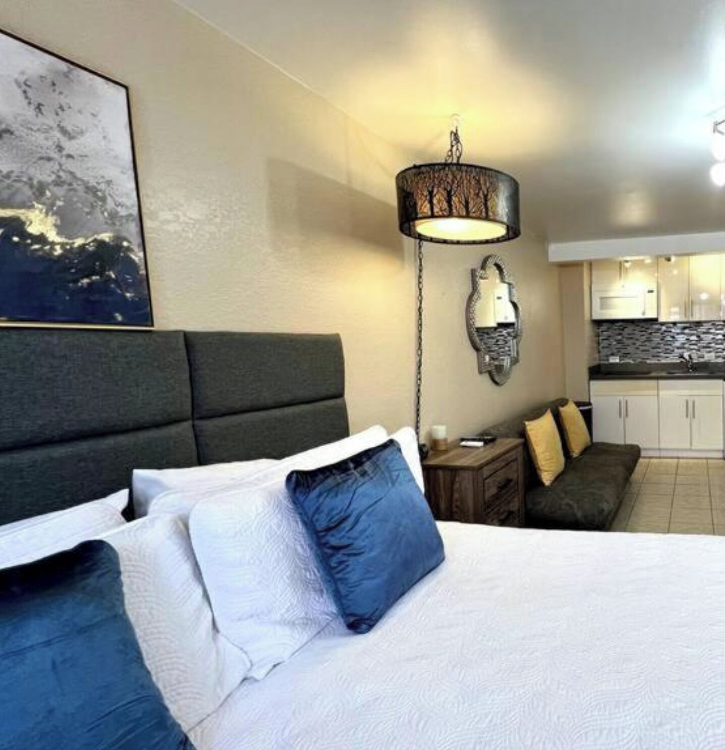 Habitación de hotel con cama, mesita de noche, lámpara, luz empotrada en la pared, un cuadro y una kitchenette al fondo.