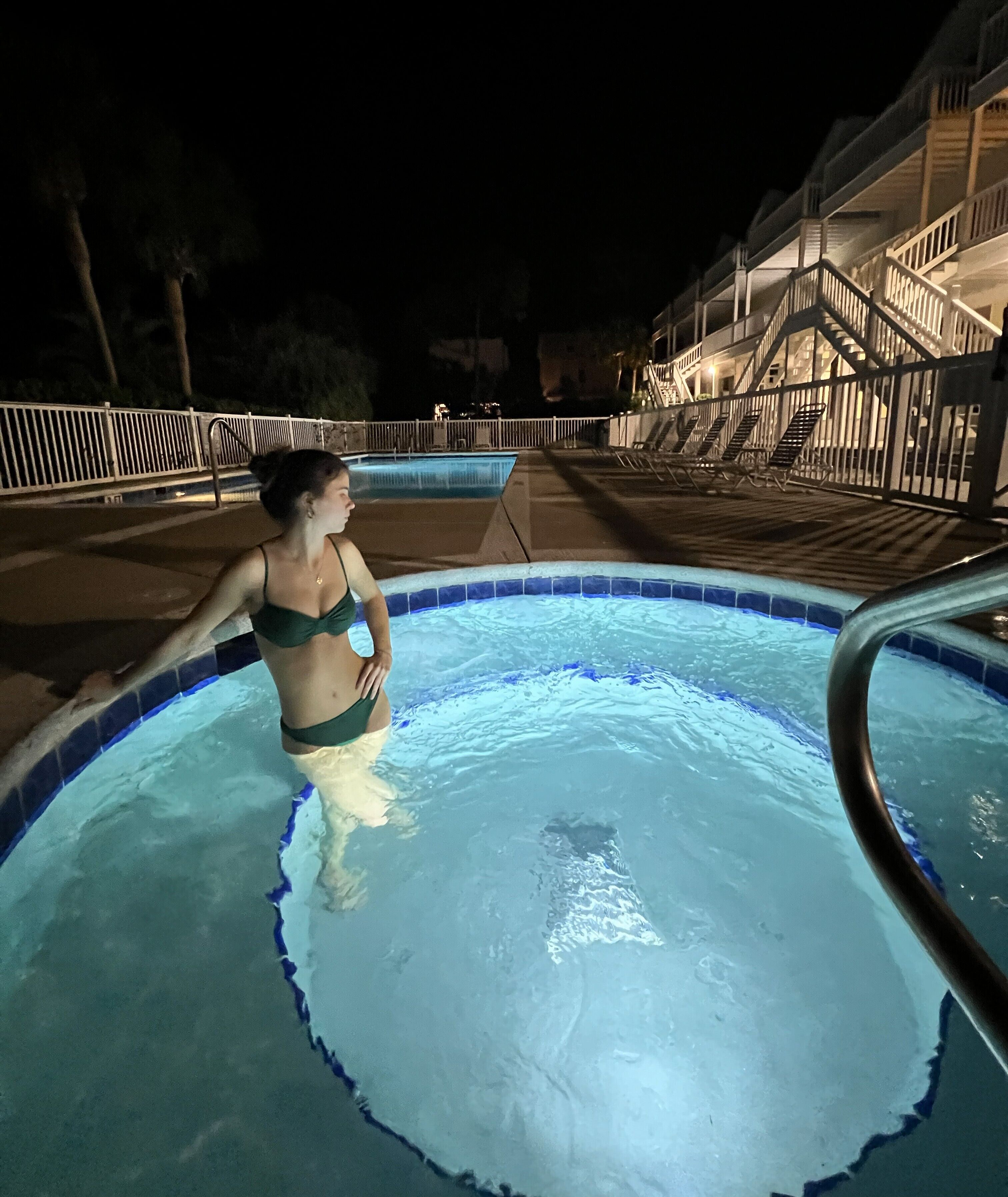 A swimming pool with a person in a bikini, a wooden deck, and a staircase leading to an upper level.