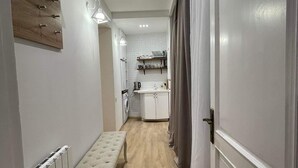 Un couloir étroit avec un banc, un radiateur et une porte donnant sur un coin cuisine équipé d’un plan de travail et de placards.