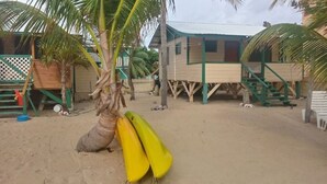 Una zona arenosa con una palmera, una estructura de madera con techo verde y un pequeño edificio blanco.