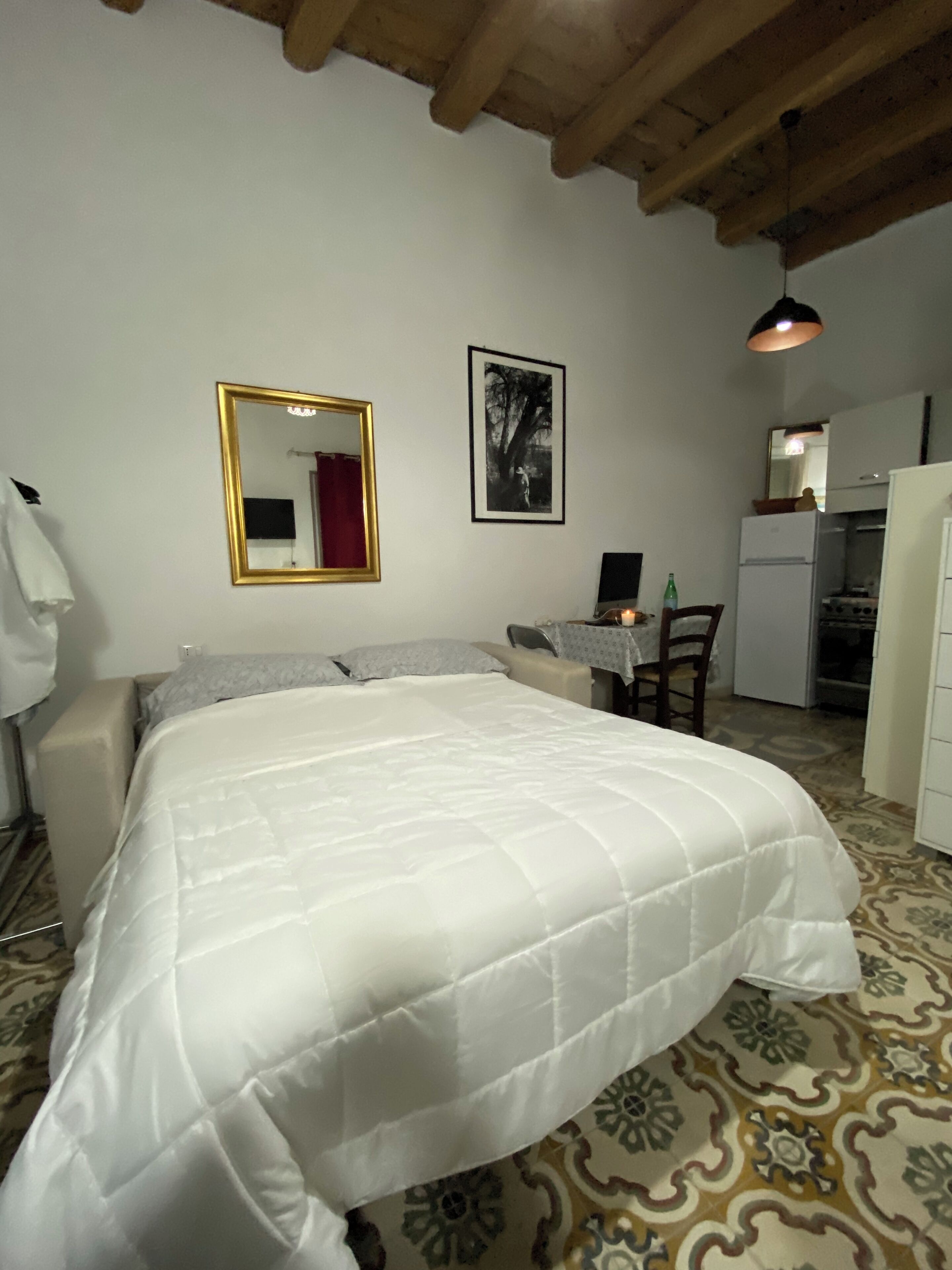 A bedroom with a bed, a desk, a chair, a mirror, and a refrigerator.