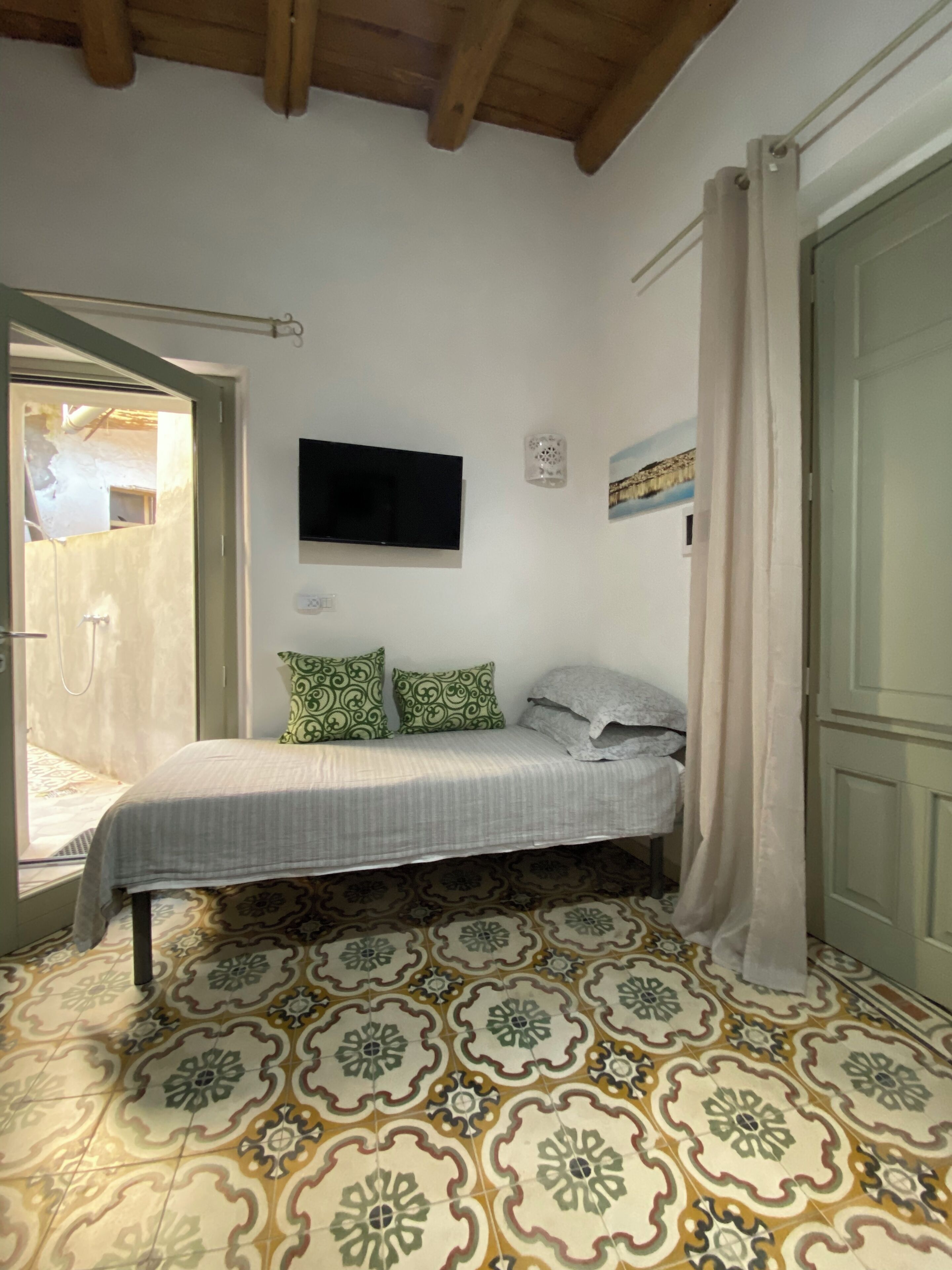 A bedroom with a bed, a television mounted on the wall, patterned floor tiles, and a door.