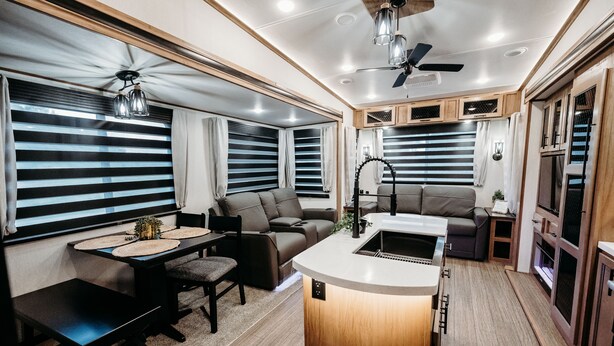 Un intérieur de camping-car compact comprenant un coin cuisine, un espace salon et de grandes fenêtres équipées de stores.