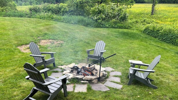 Ein Lagerfeuer, umringt von Adirondack-Stühlen, in einer grünen Außenanlage.