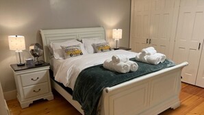 Ein ordentlich bezogenes Bett mit weißer Bettwäsche und einer grünen Tagesdecke, zwei Nachttischlampen sowie ein Nachttisch mit Spiegel und Büchern.