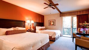 Quarto de hotel com duas camas, uma mesa, uma cadeira, ventilador de teto e varanda com cortinas.