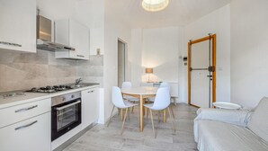 Eine moderne Küche mit weißen Schränken, einem Gasherd und einem Backofen. Es gibt eine Essecke mit einem runden Tisch und weißen Stühlen. Ein Sofa und eine Lampe sind ebenfalls zu sehen.