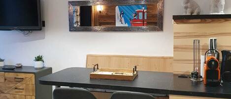 Une cuisine moderne avec un îlot central en bois, deux tabourets de bar noirs, un téléviseur à écran plat fixé au mur et une horloge rouge et blanche ornée d’un motif d’ours.