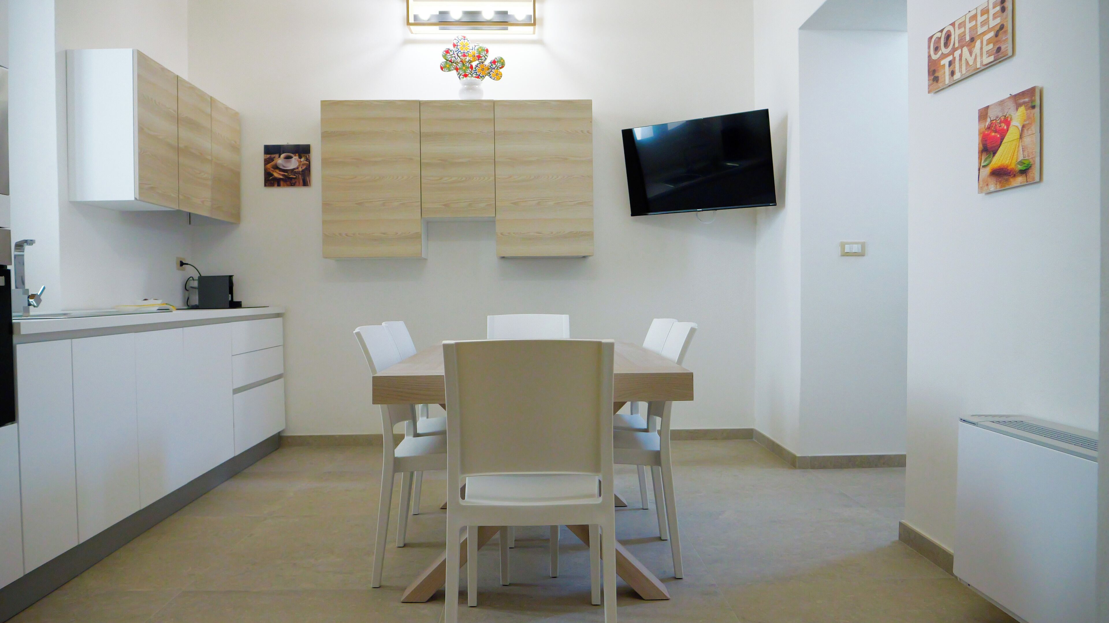 A modern kitchen with white cabinets, a wooden dining table, and chairs. A flat-screen TV is mounted on the wall above the table.
