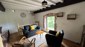 Ein geräumiges Wohnzimmer mit einer schwarzen Couch, einem Holzstuhl und einem kleinen runden Tisch. Eine Uhr ist an der Wand befestigt, und es gibt ein Fenster mit Blick auf Grünflächen.