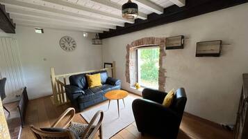 A spacious living room with a black sofa, a wooden chair, and a small round table. A clock is mounted on the wall, and there is a window with a view of greenery outside.