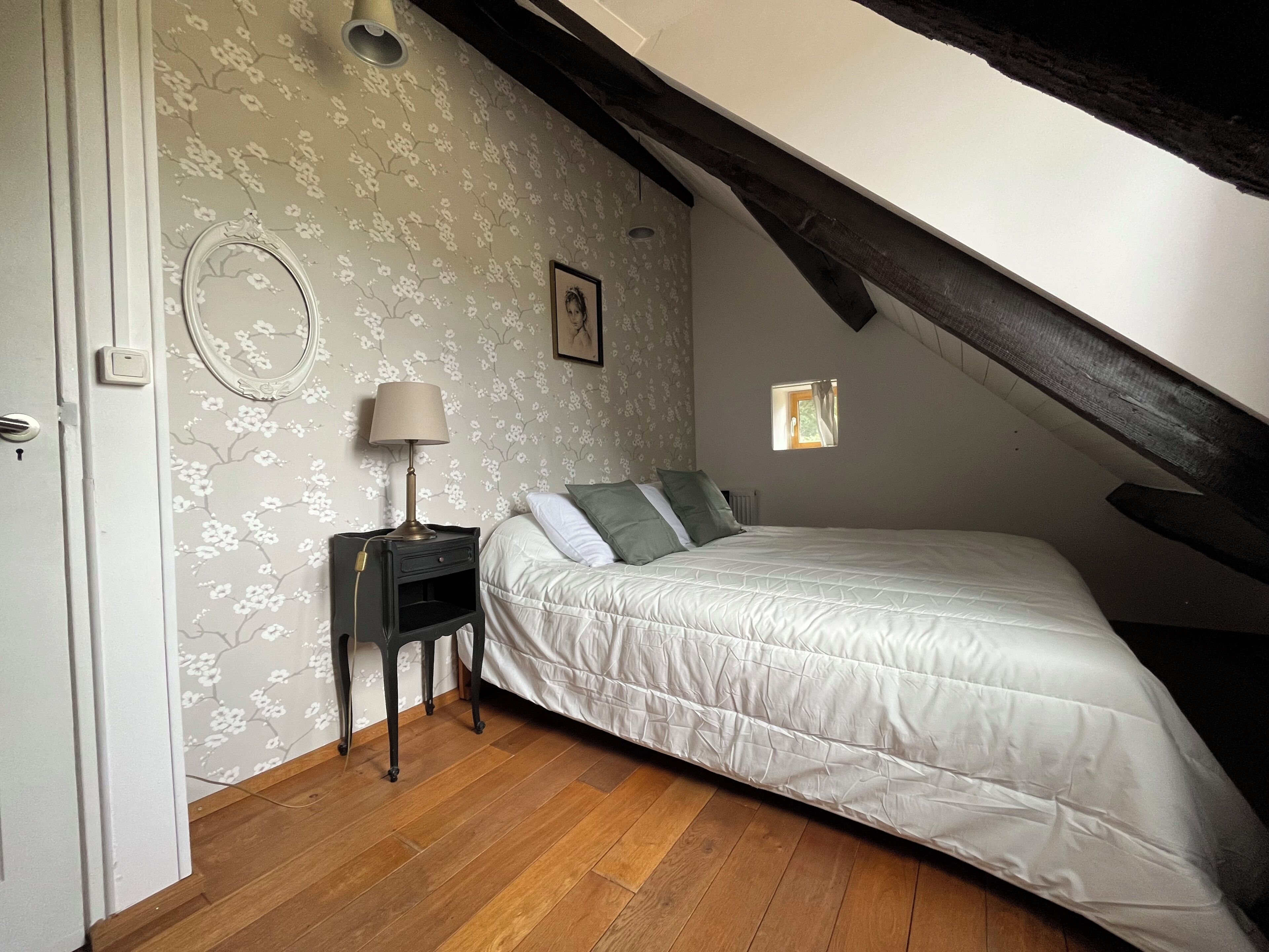 A bedroom with a sloped ceiling, a bed with white bedding, a nightstand with a lamp, and a floral wallpaper pattern.