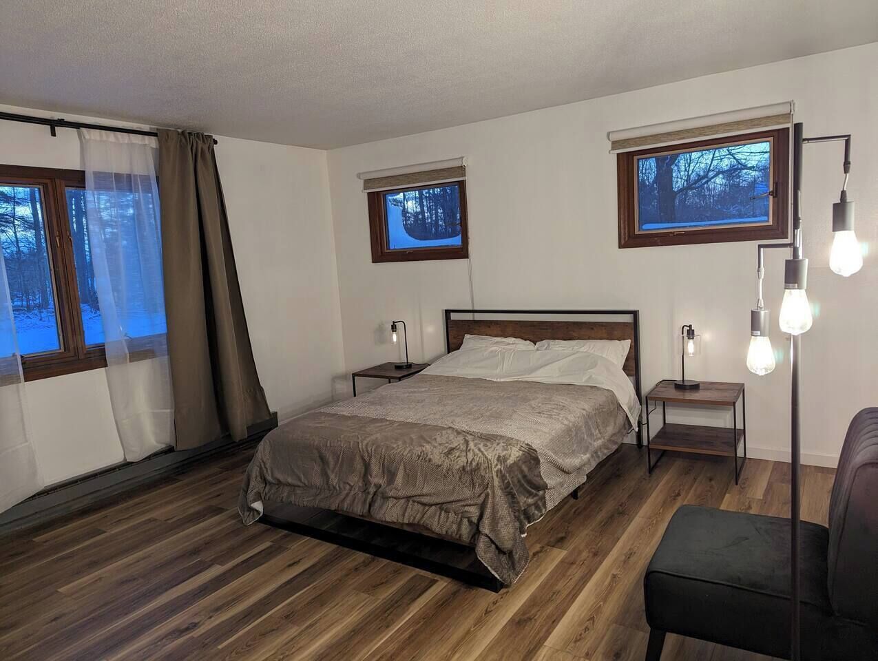 Un dormitorio con una cama, dos mesitas de noche, una silla y dos ventanas con cortinas.