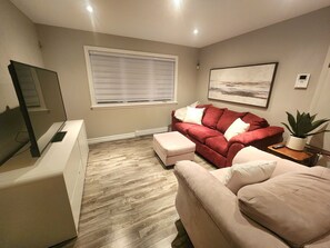 Una sala de estar con un sofá rojo, un sofá beige, un televisor, una ventana con persianas, un cuadro en la pared y una planta en maceta.