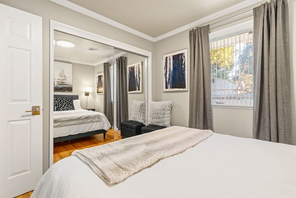 Quarto de hotel com uma cama grande, uma cama menor, um sofá e uma escrivaninha. Há uma janela com cortinas, um quadro na parede e uma porta.