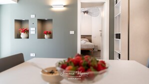A modern dining area with a table, chairs, and a bowl of fruit. There are wall-mounted light fixtures and a door leading to a bedroom. - Cozy and uniquely designed apartment (Riva di Solto)